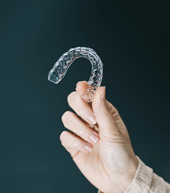 Close-up of a hand holding a transparent dental aligner used for teeth straightening against a dark background.
