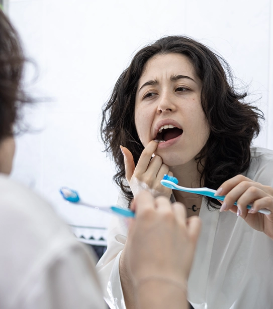 Young woman looking in the mirror and touching her tooth while holding a toothbrush, showing signs of dental pain.
