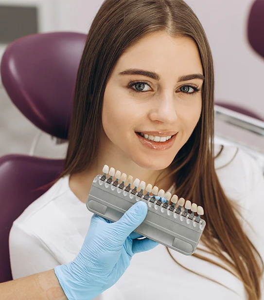 A female dentist in blue gloves holding a professional shade guide against a patient's smile to select the perfect color for a dental implant crown.