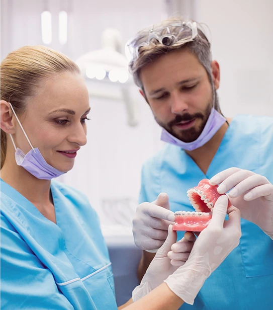Two dental professionals in blue scrubs and gloves examining a dental model with metal braces in a clinical setting.