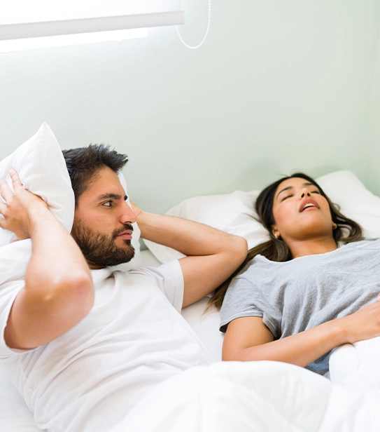 man covering ears with pillow due to partner snoring loudly in bed